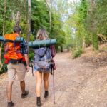 Two backpackers hiking along a wooded trail towards their campsite.