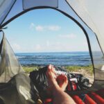 A backpacker sitting inside a large camping tent located on a beach with the ocean in the background.