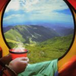 A backpacker sitting inside a two-person camping tent located on a mountain top with a view of a green mountain valley in the background.