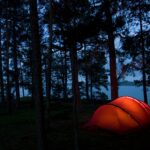 Two person camping tent illuminated beautifully in the night at a wooded campsite near a mountain lake.